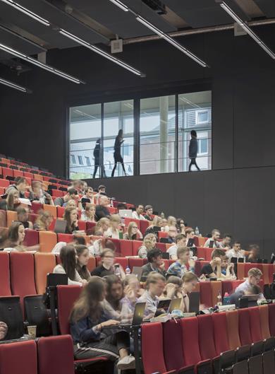 Studierende sitzen in einem großen Hörsaal mit roten und orangefarbenen Sitzen. Viele arbeiten an Laptops oder machen Notizen; im Hintergrund sind durch ein großes Fenster Personen auf einem Flur zu sehen.